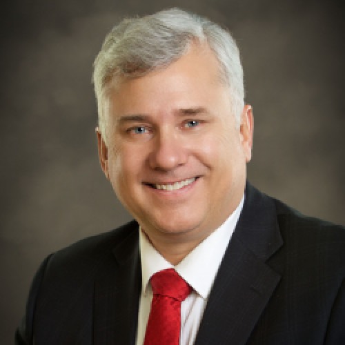 Headshot of Karl wearing a black suite and red tie against a gray-brown backdrop